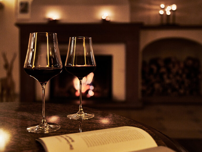 Der Kamin in der Bar & Lounge im Hotel Kronelamm sorgt für gemütliche Stunden an kalten Wintertagen. Zwei Gläser Rotwein stehen auf einem Tisch vor einem angefeuerten Kamin