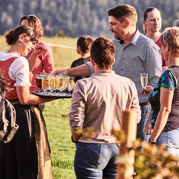 A young service employee holds a tray with champagne glasses in her hand and distributes them to the guests standing around.