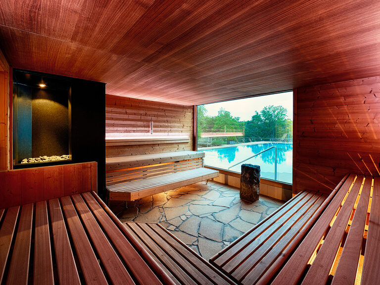 Rotbeleuchtete Sauna mit Sitzgelegenheiten aus Holz und großem Panoramafenster mit Blick auf den Außenpool.