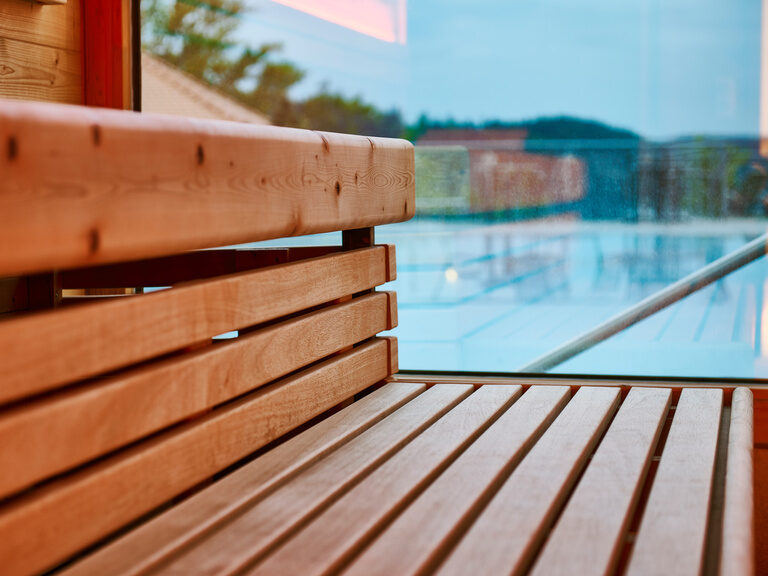 Sitzbänke in einer Sauna, die durch ein Fenster vom Pool getrennt sind.